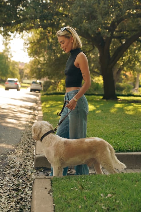 Blonde Woman Wearing A Black Top And Jeans Walks Dog In The Street In The Neighborhood With Green Lawn