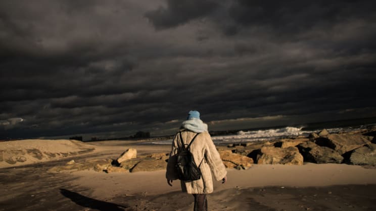 A Woman Wears A Black Backpack While Walking Along An Ocean Coast At Sunset
