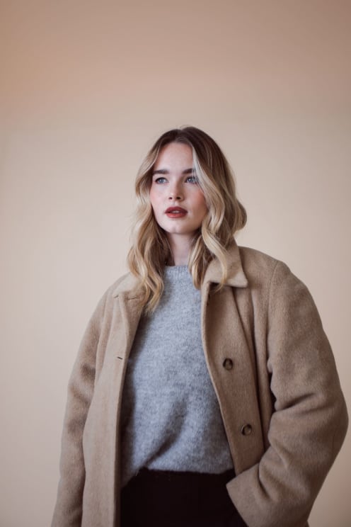 Woman Wearing A Beige Coat Over A Gray Sweater, Standing Against A Light Background
