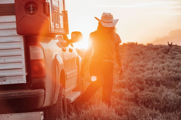 Woman Wearing A Cowboy Hat Stands Next To A Truck As The Sun Shines Brightly On The Horizon