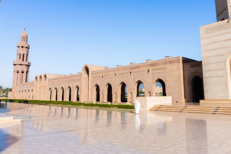 A Person In White Robes Walks Along The Exterior Of A Large Mosque With A Tall Minaret, A Series Of Arches, And A Vast Courtyard Paved In Light-colored Stone