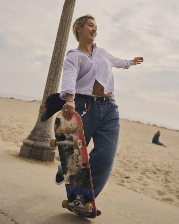 Young Woman Smiles As She Holds Her Skateboard On A Sidewalk At The Beach