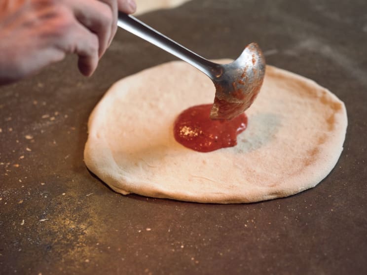 Hand Holding A Metal Ladle, Spreading Tomato Sauce Over A Raw Pizza Dough