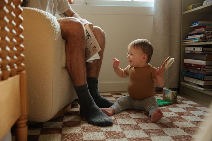 Baby Sitting On Rug Next To Armchair With Adult Person
