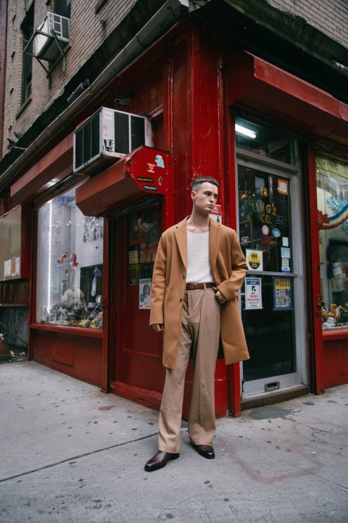 A Serious Looking Young Man With A Cropped Hair Cut, Standing Outside A Corner Shop