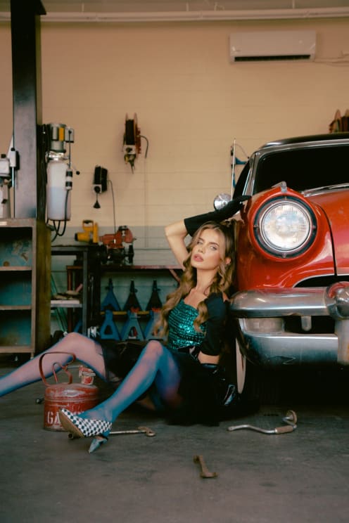 Young Blonde Woman In Dress And Skirt Sitting In A Workshop Next To A Red Classic Car