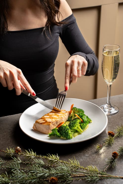 Woman In Black Top Cuts Grilled Salmon With Steamed Broccoli On A White Plate Next To A Glass Of Champagne On A Decorated Table