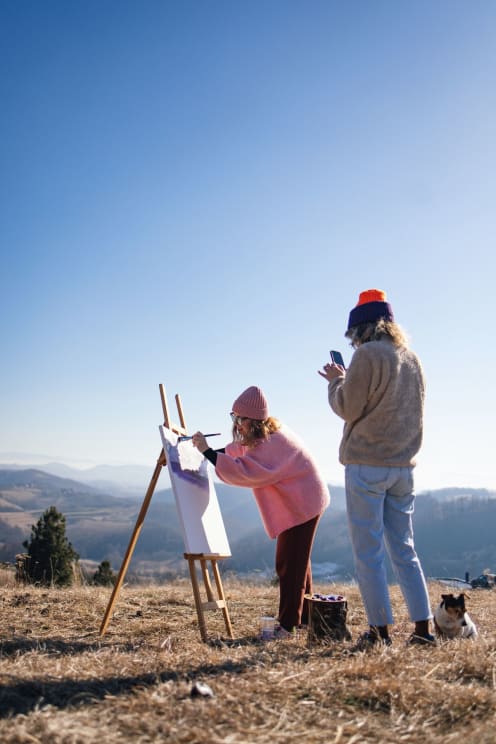 Woman Photographing Another Woman Who Is Painting A Picture On A Hill With Mountains In The Background