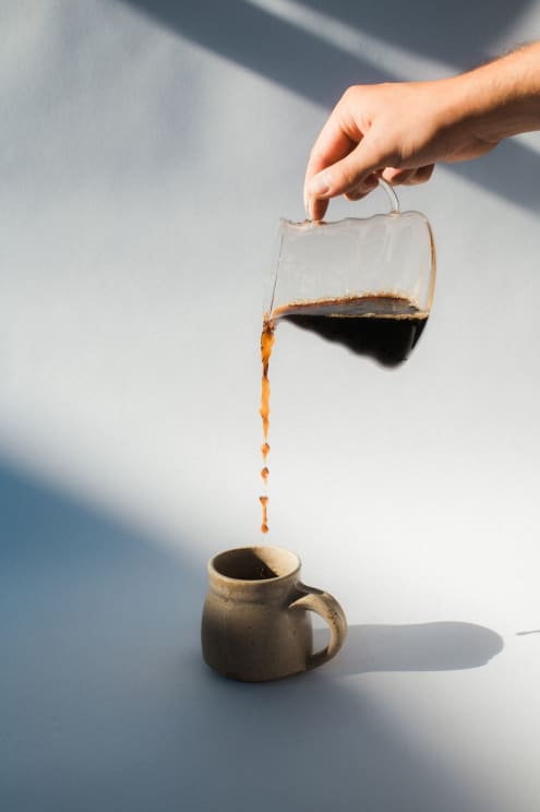 A Hand Pours Coffee From A Glass Carafe Into A Ceramic Mug