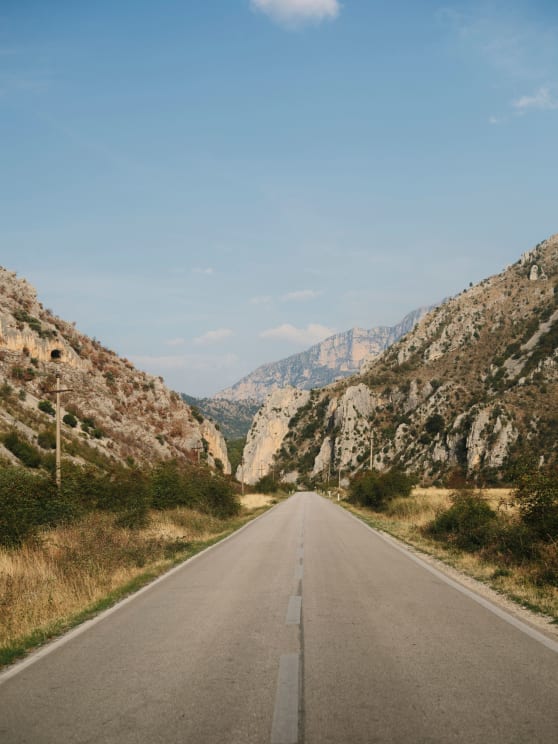 Straight Road Stretching Through A Rocky Valley With Dry Vegetation, Flanked By Rugged Hills Under A Clear Blue Sky