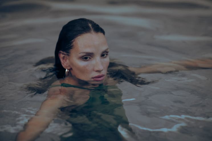 A Woman With Wet Hair And Hoop Earrings Floats In Calm Water, Wearing A Dark Green Swimsuit