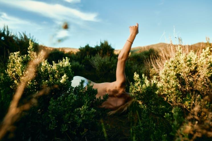 A Person Lies Among Vibrant Green Shrubs Under A Bright Blue Sky, Their Arm Raised As If Reaching Or Gesturing, With Hills Visible In The Distance
