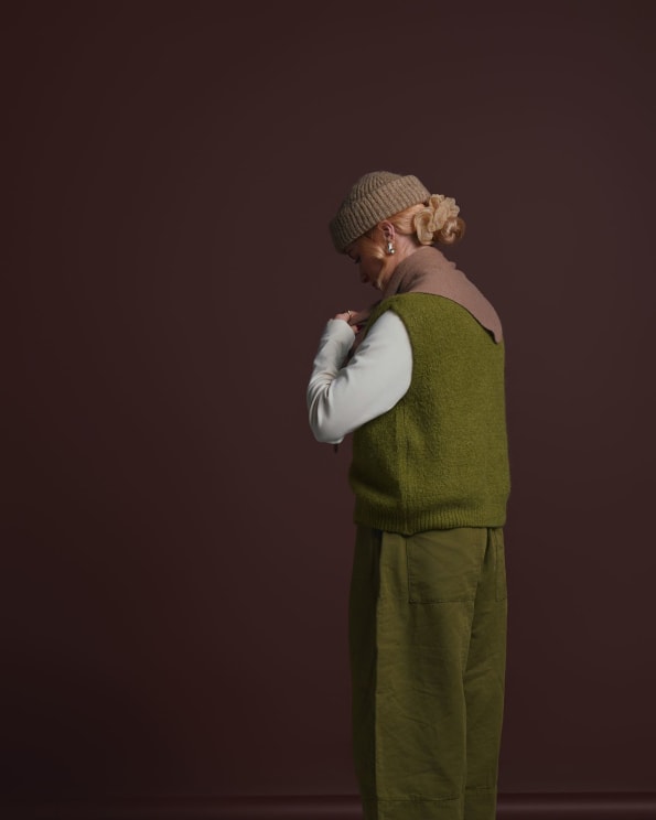 Woman Standing In A Room Wearing A Knit Cap And Sweater Vest Over A Shirt With Baggy Pants