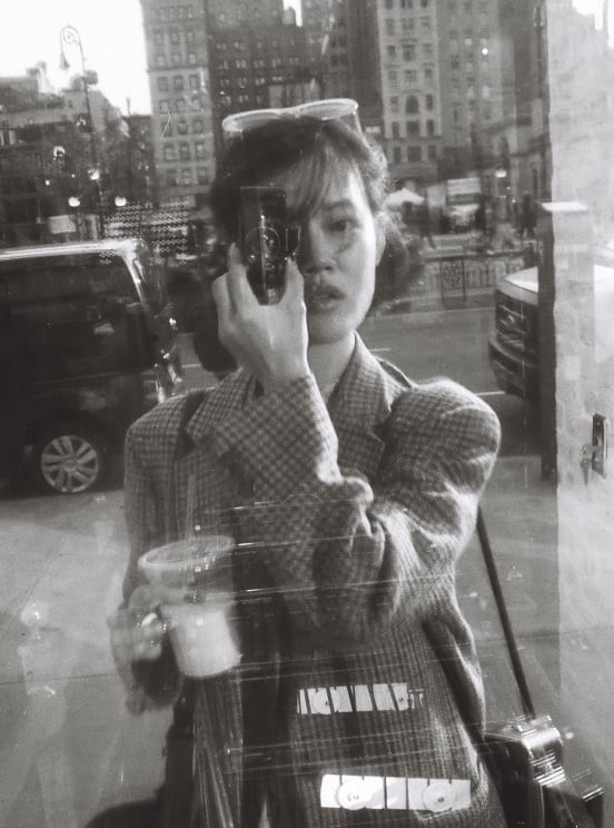 Woman Standing On A Downtown Street Holding Her Camera Up To Her Face And A Drink In The Other Hand