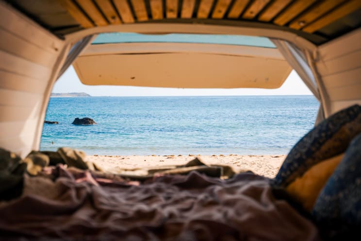 View From Inside A Van With The Back Door Open, Revealing A Tranquil Beach With Golden Sand, Crystal Blue Sea And Rocks In The Distance