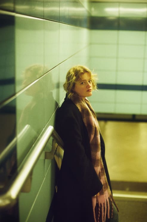Blonde Woman Leaning Against A Green Tiled Wall In A Subway Station