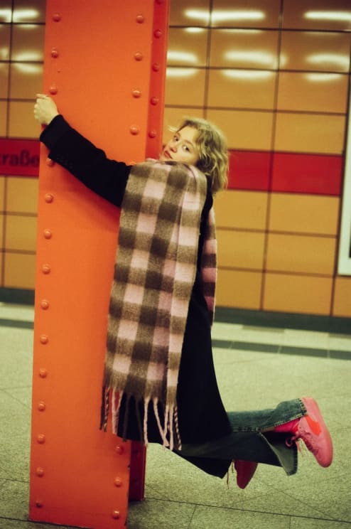 Portrait Of A Young Blonde Woman Who Has A Beige Scarf Around Her Neck And She Is Hanging From The Support Beam Of An Orange Terminal
