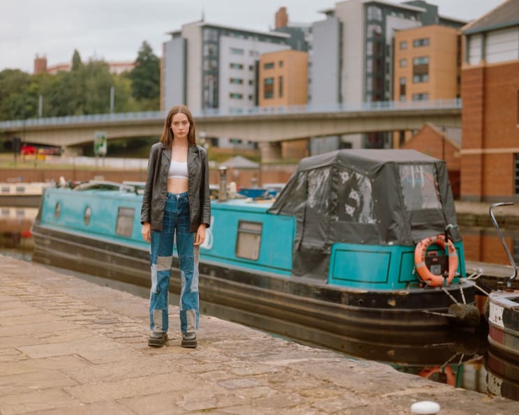 A Young Woman In Front Of A Blue Boat On An Urban Canal With Modern Buildings In The Background