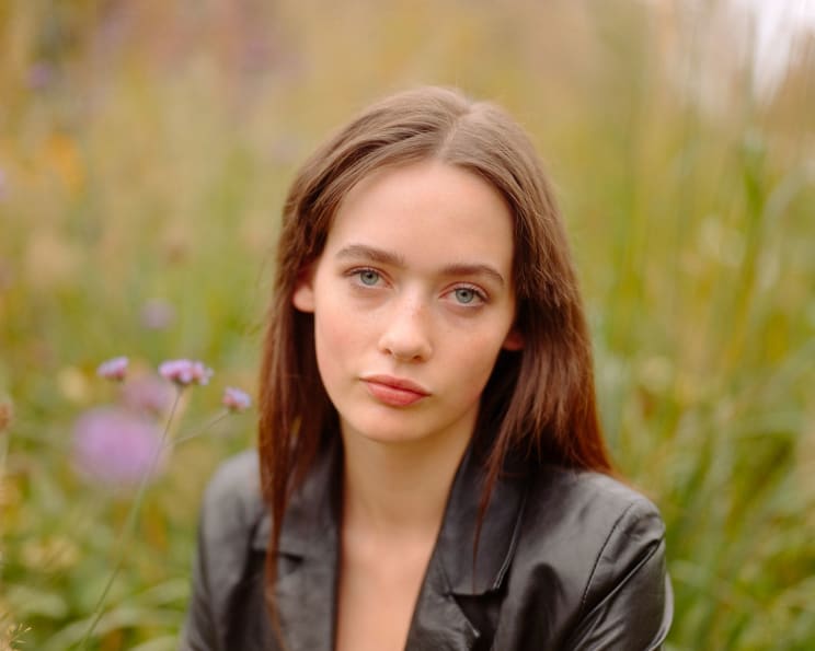 A Fair-skinned Young Woman With Long, Brown Hair And Blue Eyes, Wearing A Black Leather Jacket, Poses In Front Of A Blurred, Flowery Meadow