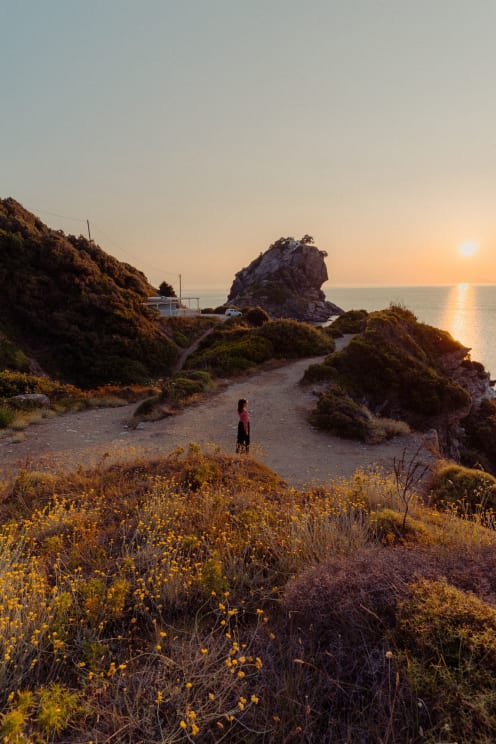 Woman Is Standing On A Path Surrounded By Vegetation And Small Yellow Flowers, With A Rock In The Sea In The Background And The Sun Setting On The Horizon