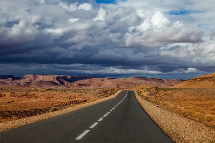 Deserted Asphalt Road Advances Through An Arid Landscape, Surrounded By Rocky Mountains Under A Sky Filled With Dense Clouds