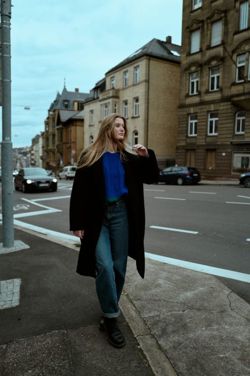 Woman Wearing A Black Coat, Blue Sweatshirt And Jeans Walking Down A Street With Historic Buildings And Cars In The Background