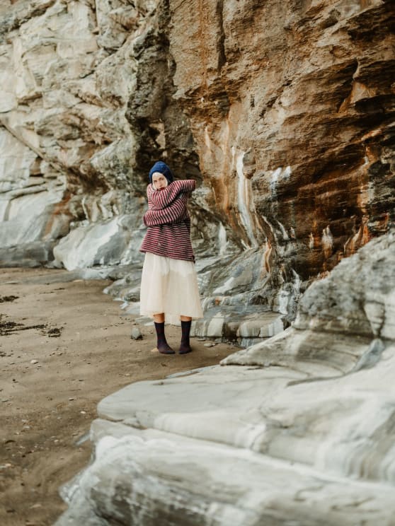 Woman Huddled Against A Towering Rock Wall, Dressed In Cozy Clothing
