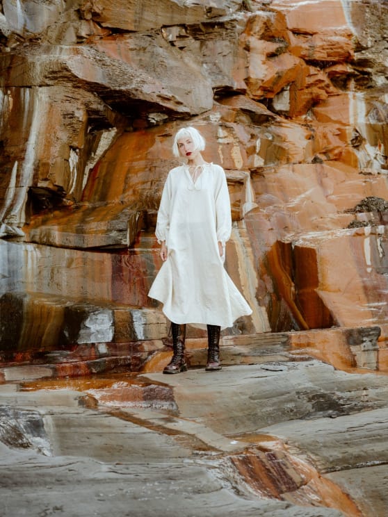 Woman In A Long White Dress Wearing Leather Boots Standing In A Natural Environment Surrounded By A Stone Wall
