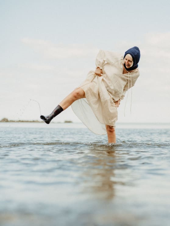 Woman In A Light Dress And Blue Hat Plays In The Water, Lifting Her Leg And Splashing Drops