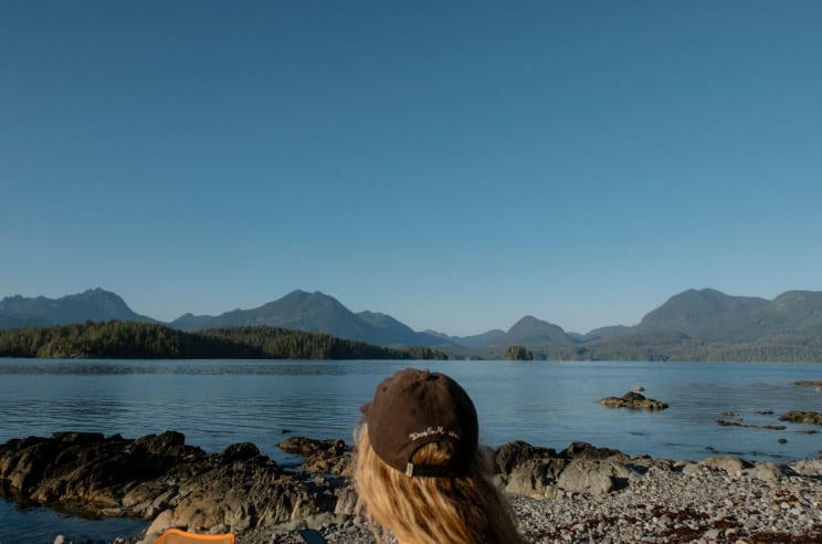 Woman Wearing A Cap Sitting Along The Coast Looking At Mountains Across The Water
