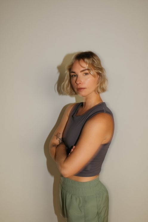 A Short-haired Blonde Woman Wearing A Sleeveless Gray Top And Green Pants Stands Confidently With Arms Crossed, Displaying A Bird Tattoo And A Flower Tattoo On Her Arm Against A Neutral Background