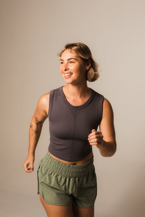 Woman With Short Blonde Hair, Wearing A Gray Top And Green Shorts, Moves With Her Arms Flexed