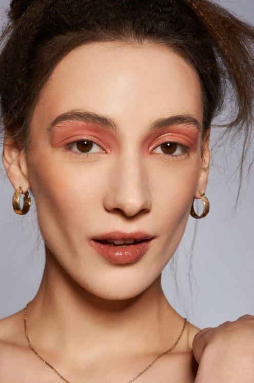 A Woman Wearing Her Hair Tied Above Her Head, Heavy Eye Make Up, Earrings, A Thin Necklace, And A Tiny Smile