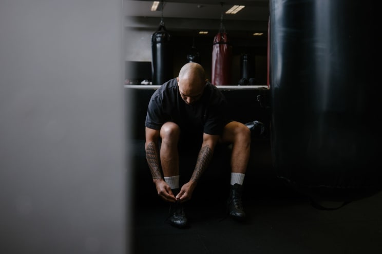A Bald Man With Geometric Tattoos On His Legs Ties His Shoes In A Boxing Gym, Wearing Black Workout Clothes, While Punching Bags Hang In The Background