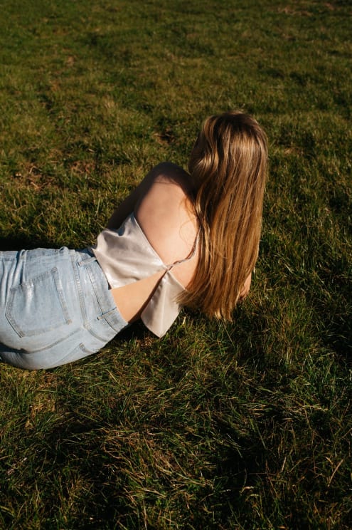 Woman With Long Blonde Hair Lies On Green Grass Wearing A Satin Top And Light Blue Jeans In Sunlight