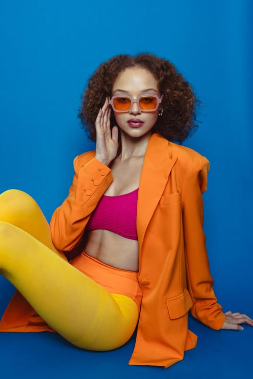 A Curly-haired Woman Wearing Tinted Orange Sunglasses, A Bold Orange Blazer, A Bright Pink Top, And Yellow Tights Sits Confidently Against A Vibrant Blue Backdrop