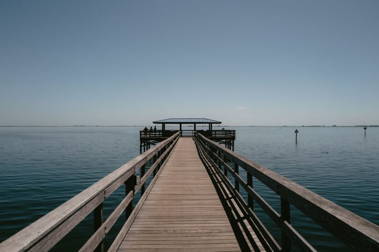 A Long Wooden Pier Stretches Over Calm Waters Towards A Covered Kiosk