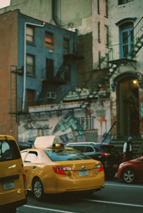 In A Grainy Film Shot, A Yellow Taxi Rolls Down A Busy City Street In Front Of A Weathered Building With A Mural And Fire Escapes, Capturing An Urban, Vintage Vibe