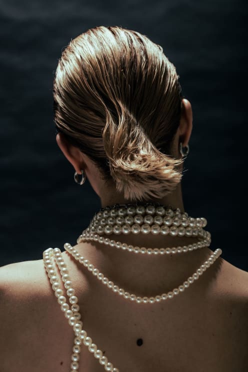A Woman Seen From The Back, With Her Hair Tied Up And Pearl Necklaces On Her Illuminated Skin