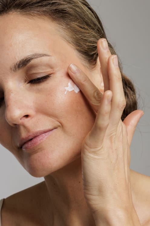 A Woman Rubs Skin Care Product On Her Cheek While Smiling