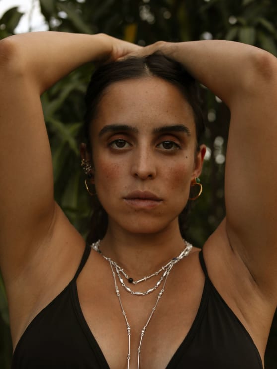 Framed Closely, A Woman With Dark Hair And Silver Necklaces Raises Her Arms Behind Her Head And Gazes Intently At The Camera, With Soft Green Foliage Behind