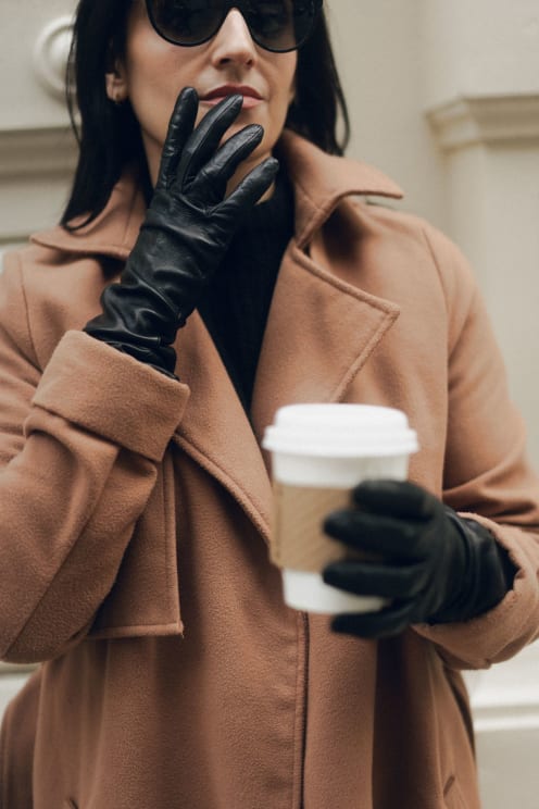 Woman Wearing An Elegant Beige Coat And Black Gloves Holds A Cup Of Coffee While Touching Her Lips Lightly With Her Gloved Hand