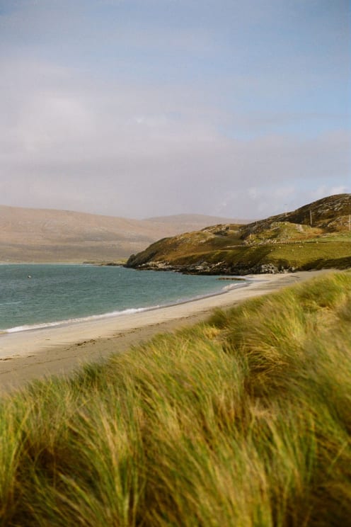 Secluded Beach Of White Sand Meets The Blue Sea, Surrounded By Dunes Covered In Vegetation And Gentle Hills In The Background