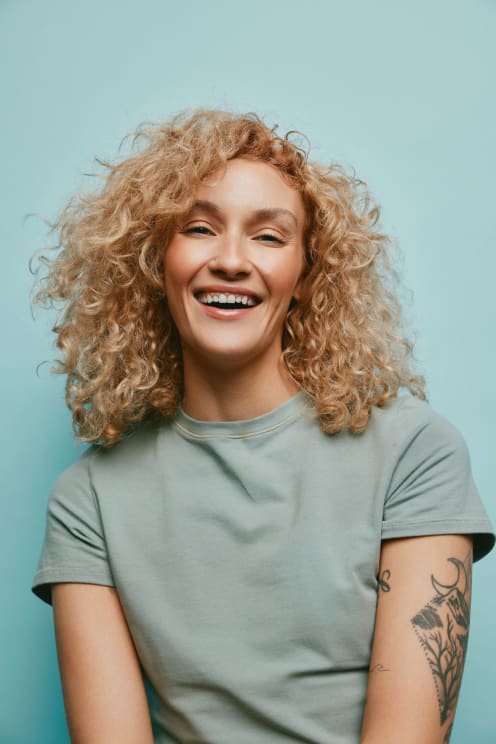 Blonde Woman With Curly Hair Wearing A Light Green T-shirt Smiles Brightly Against A Light Blue Background, Showcasing A Tattoo On Her Arm