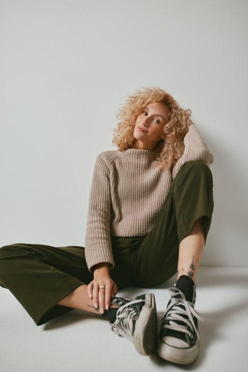 A Stylish Young Woman With Curly Blonde Hair Sitting Casually Against A Plain White Wall, Wearing A Cozy Beige Sweater, Olive Green Pants, And Worn Black Converse Sneakers, Exuding A Relaxed And Confident Vibe