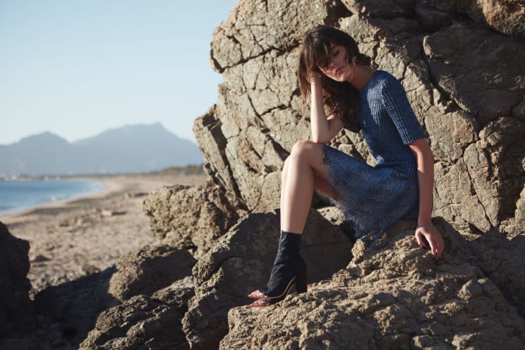 Woman Wearing A Dress Sits On Rocks On Along The Coast Near The Beach
