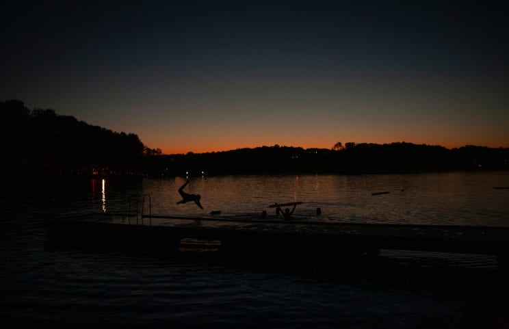 Silhouettes Of People Enjoy A Lake At Sunset. One Person Dives Off A Pier While Others Swim Nearby. The Fading Sunlight Casts A Warm Glow, With Distant Lights Reflecting On The Water