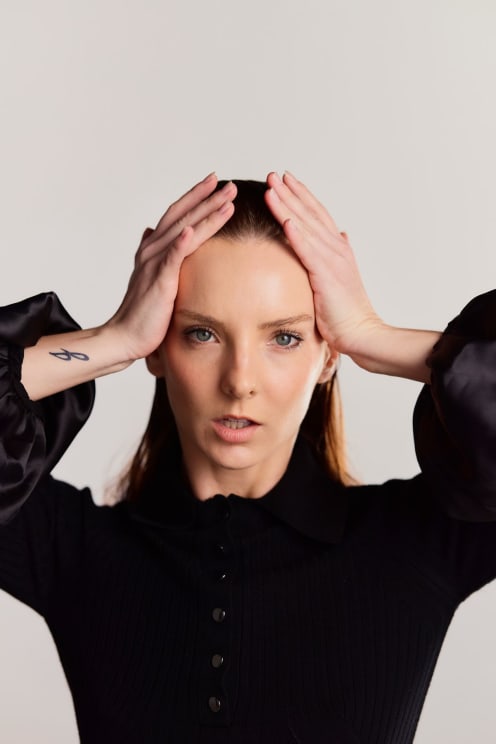 A Woman With Dark Hair And A Tattoo On Her Arm, Holding Her Head With Both Hands, Wearing A Black Shirt