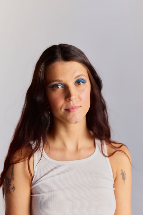 A Woman With Long Brown Hair, Tattoos On Her Upper Arms, Wearing A Tank Top, And Blue Eye Shadow, Smiles Slightly With A Sad Expression