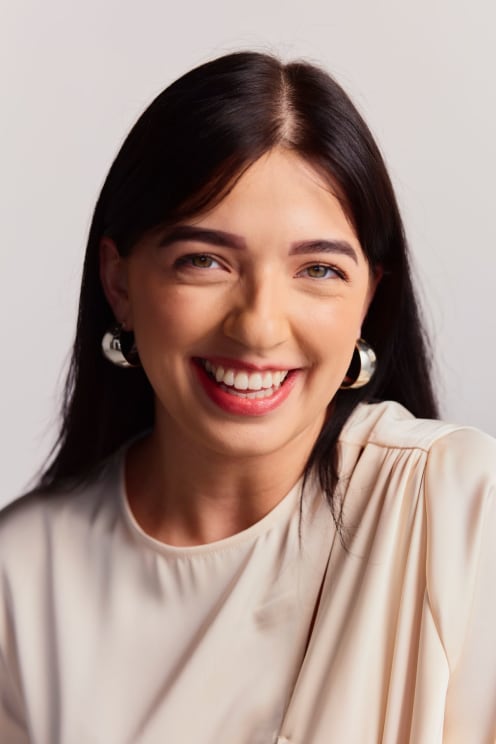 Woman Smiles Broadly, With Dark, Straight Hair, Wearing Large Earrings And A Light Blouse